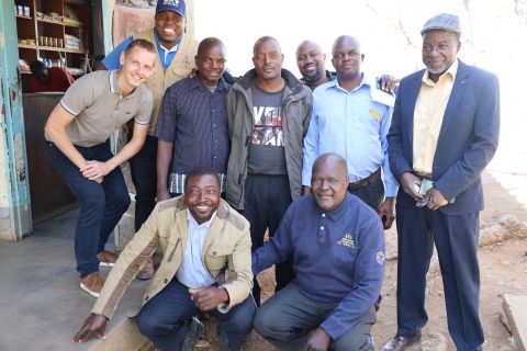 Neun Männer, darunter ein SolidarMed-Mitarbeiter, posieren fröhlich vor einem kleinen Laden in Chikombedzi.