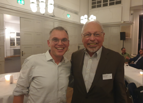 Two smiling men standing side by side in a festively lit hall.